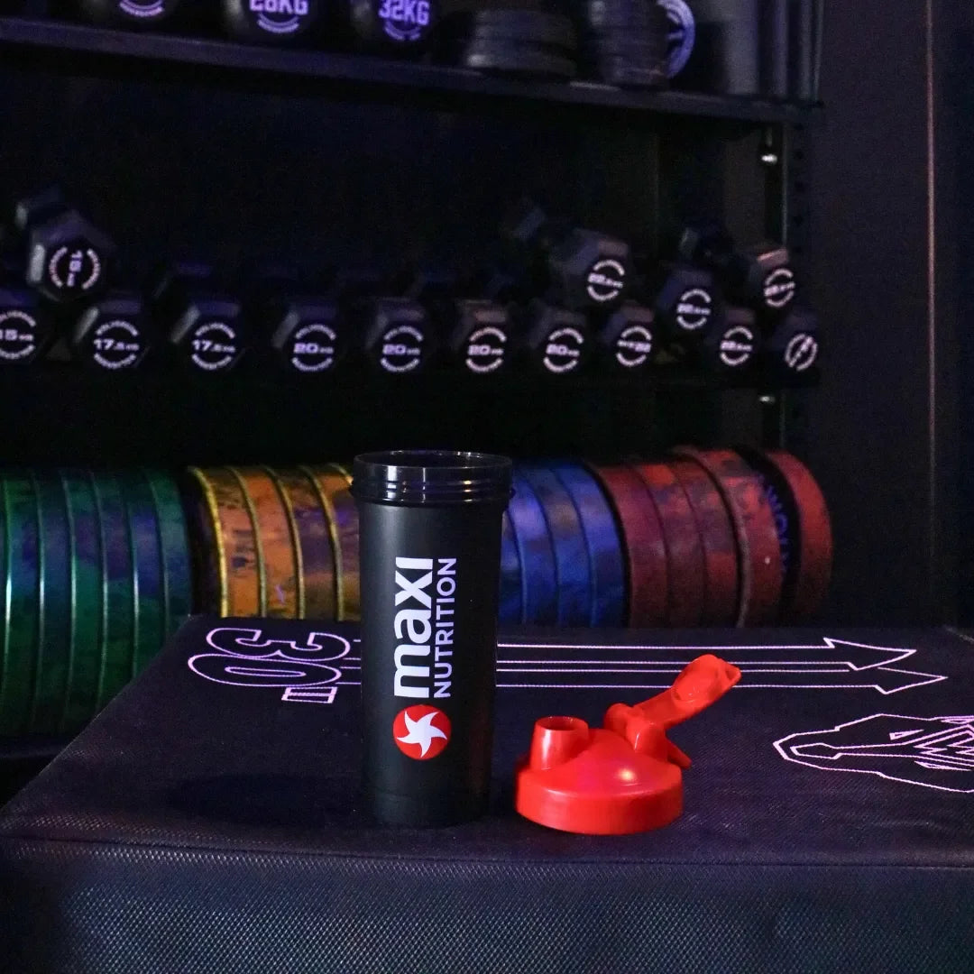 Maxi Nutrition shaker cup with a red cap on a gym floor with weights in the background